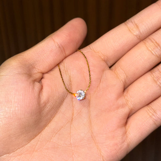 A delicate gold chain necklace with a single round clear gemstone pendant, resting on an open hand against a dark brown background.